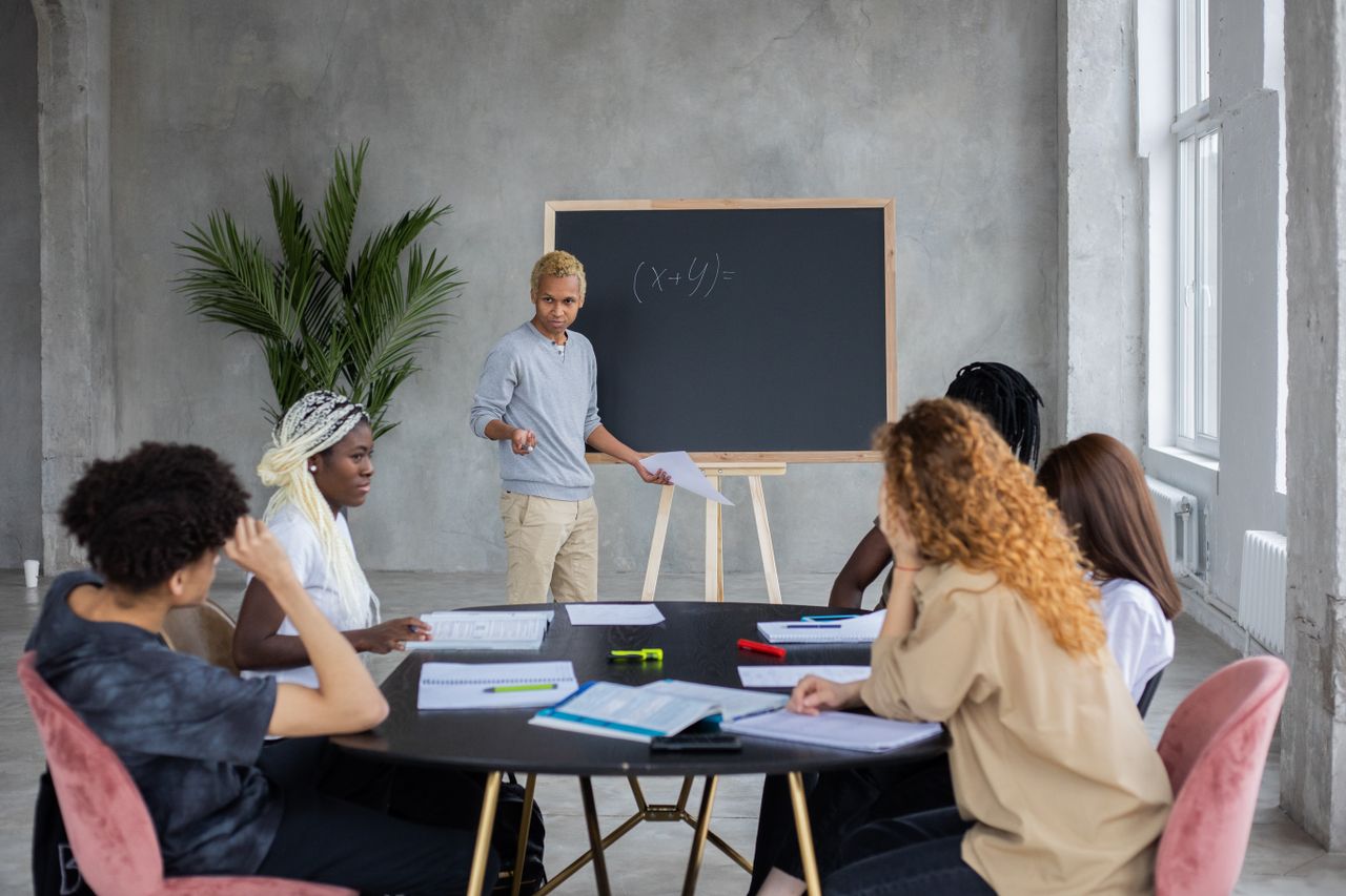 hero-img-01 African American student explaining mathematic equation to diverse classmates while doing homework together in spacious room