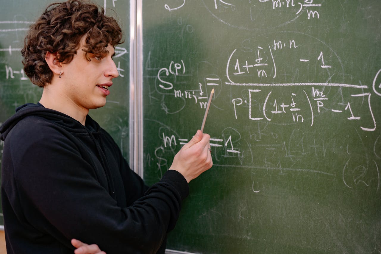 A young male educator explaining complex math equations at a university classroom blackboard.